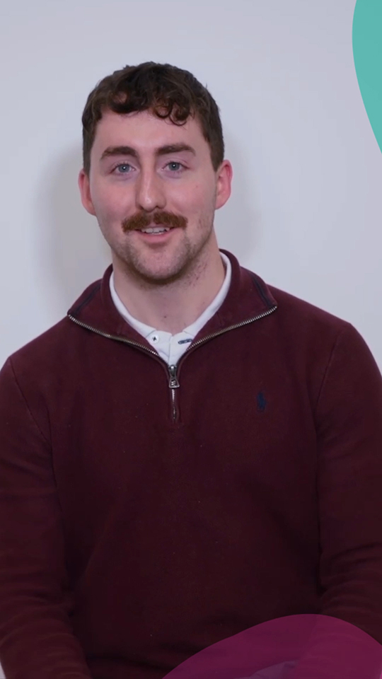 A man with short brown hair and a moustache, wearing a maroon quarter-zip jumper over a white shirt, sits and smiles in front of a plain light background with teal and maroon shapes in the corners.