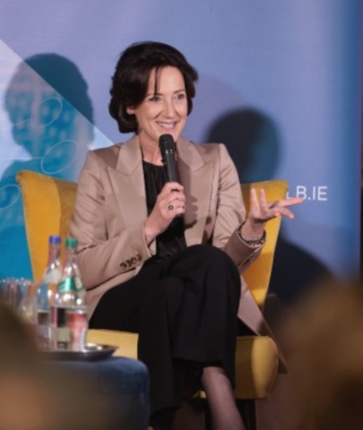 A woman with short dark hair, wearing a beige blazer and black trousers, sits in a yellow chair whilst speaking into a microphone at an event. Bottles of water are visible on a table in the foreground.