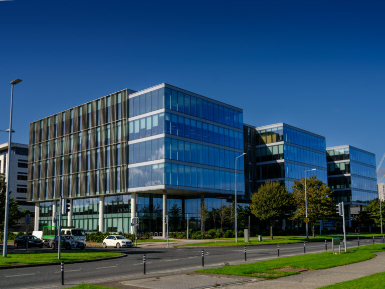 A modern office building with glass walls and multiple storeys, set against a clear blue sky. There is a green lawn, trees, and a road with streetlights and parked cars in the foreground.