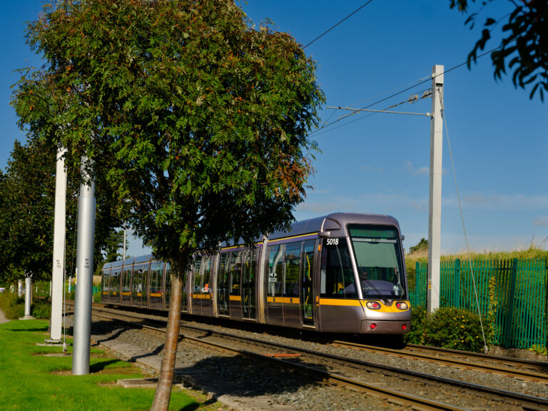 A modern light rail tram travels on tracks through a green, grassy area with trees and a fence under a clear blue sky.