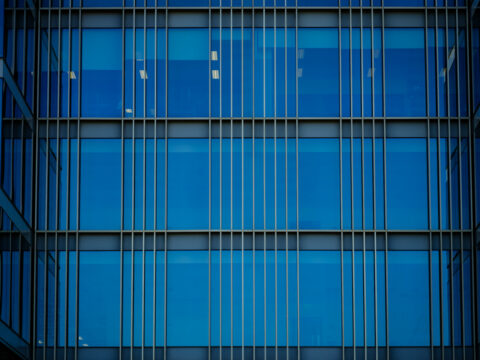 Close-up of a modern building facade with blue-tinted glass windows and vertical and horizontal metal lines creating a geometric grid pattern.
