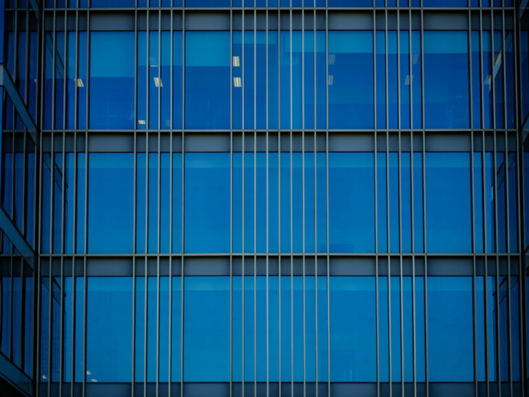 Close-up of a modern building facade with blue-tinted glass windows and vertical and horizontal metal lines creating a geometric grid pattern.