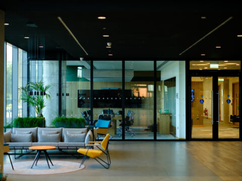 Modern office lobby with stylish seating and plants in the foreground, glass walls revealing a meeting room with chairs, a table, and a TV screen in the background. Bright natural light enters through large windows.