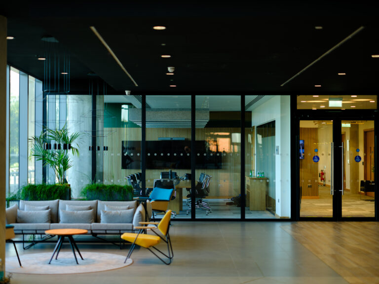 Modern office lobby with stylish seating and plants in the foreground, glass walls revealing a meeting room with chairs, a table, and a TV screen in the background. Bright natural light enters through large windows.