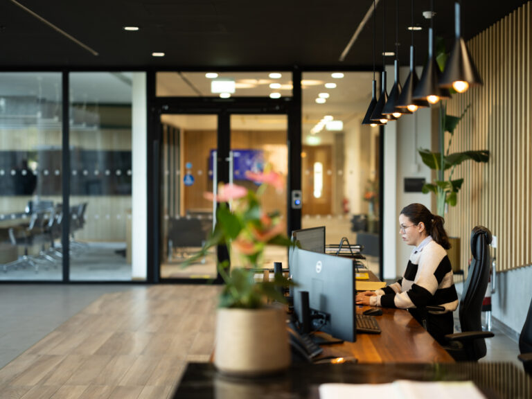 A woman wearing glasses and a striped jumper works at a computer in a modern office with wooden floors, hanging lights, and glass-walled meeting rooms in the background. A potted plant is in the foreground.