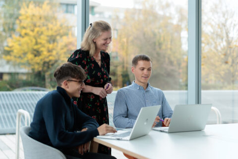 Three people are gathered around a table with laptops. Two young men are seated and typing whilst a woman stands beside them, smiling. Large windows in the background show trees with autumn foliage.